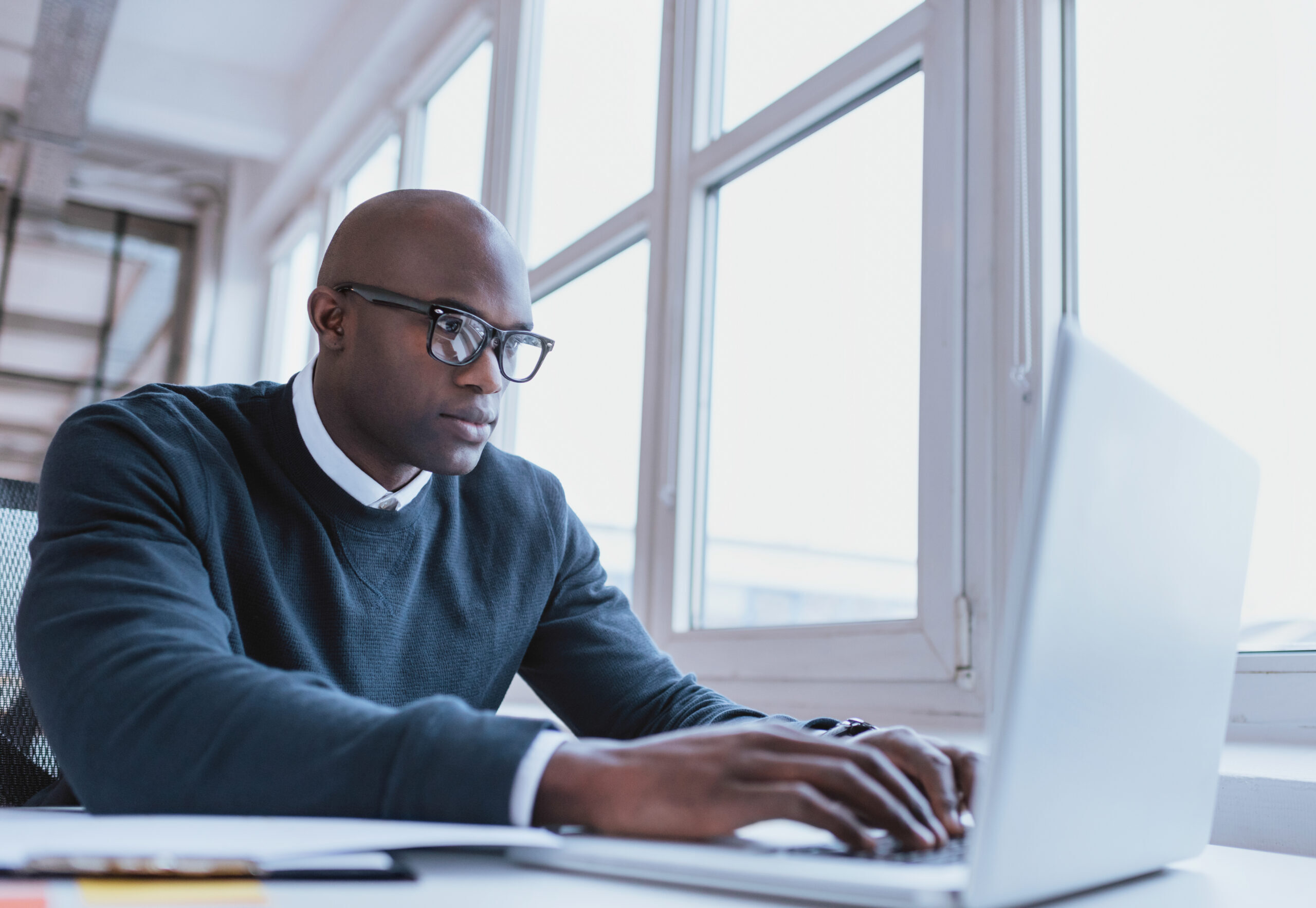 A man wearing glasses is focused on his laptop, engrossed in his work.
