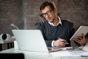 A man wearing glasses is focused on his laptop, engrossed in his work.