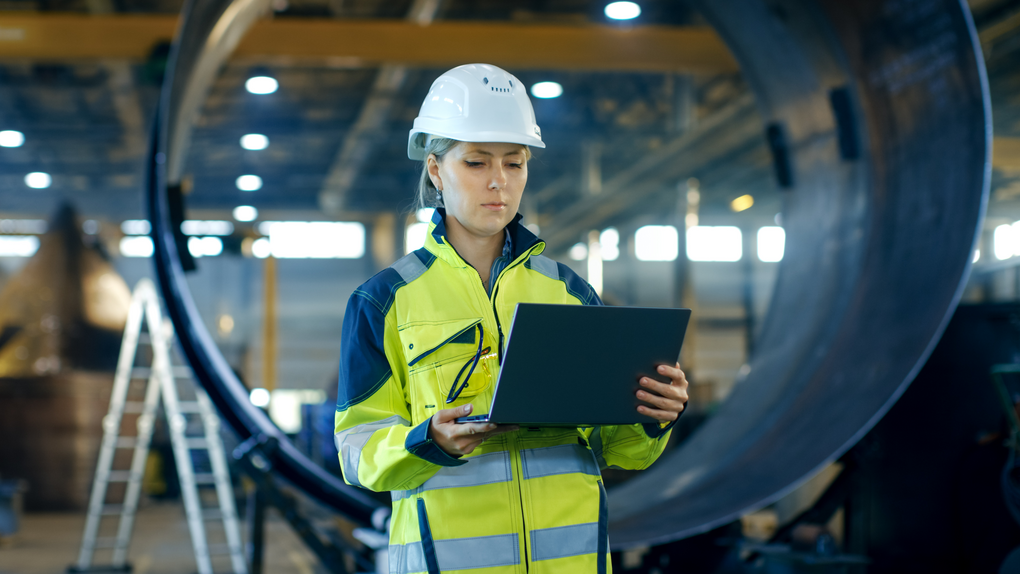 A woman in a hard hat and safety vest holding a laptop, ready to work on a construction site.